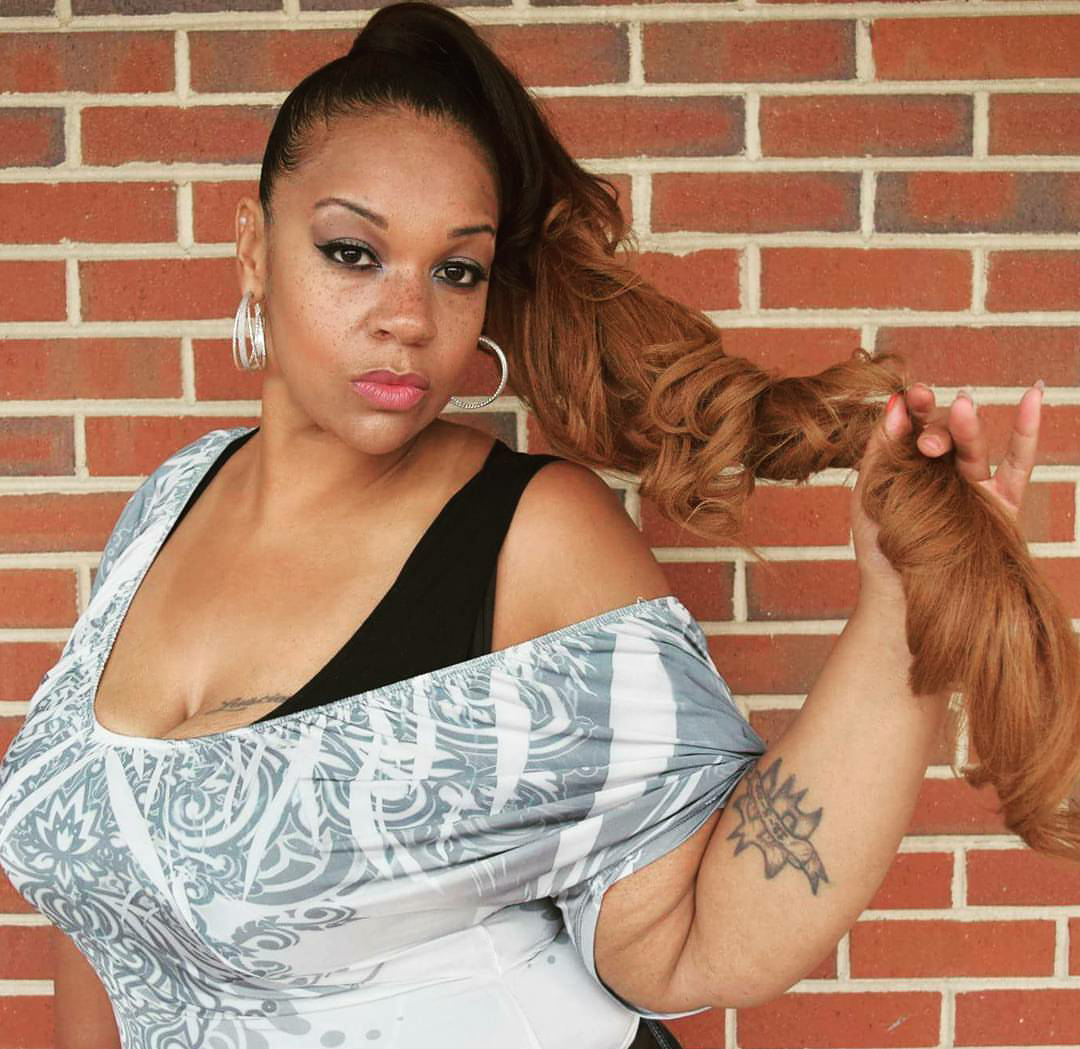 Woman posing against a brick wall, holding her long ponytail and wearing a patterned off-shoulder top.